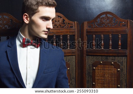 English gentleman beauty concept. Portrait of young and handsome man in blue jacket, Scottish bow-tie and white shirt posing over vintage screen. Close up. Studio shot