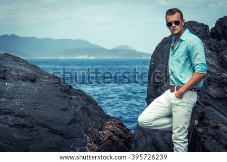 Sea/beach fashion concept. Portrait of a young and handsome man in a aqua shirt, white pants and trendy glasses posing over the shore rocks of a white sand beach. Close up.