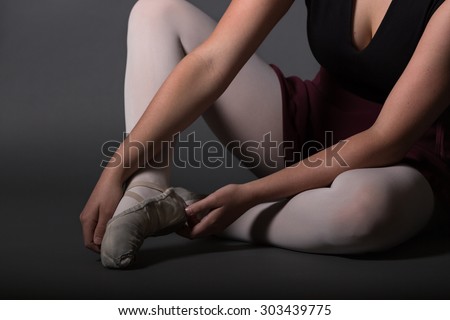 Ballet dancer in light pink tights and black leotard putting on ballet shoes in front of a dark grey background.