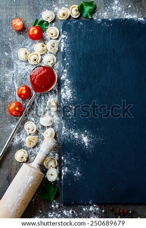 Raw homemade pasta ravioli with flour, parsley, tomatoes and vintage kitchen accessories on dark vintage background . Top view.