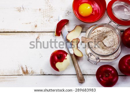 Ingredients (fresh red apples, eggs, flour in jar) on white wooden background for apple pie cooking.