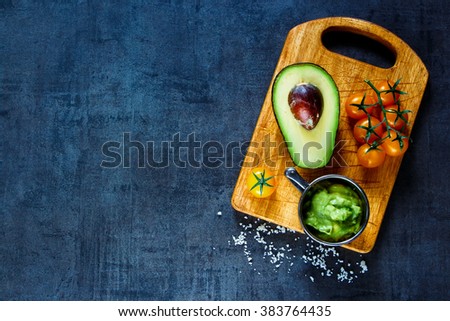 Fresh ingredients for homemade guacamole (avocado, tomato, salt) on rustic wooden cutting board over dark vintage table, top view. Healthy food background with space for text.