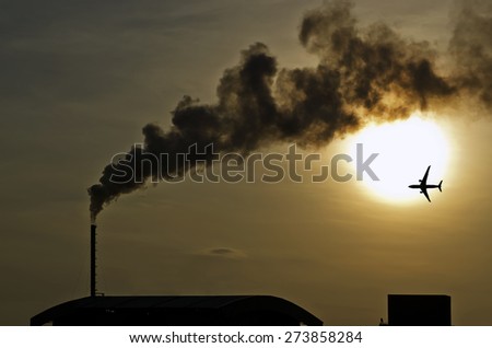 The airplane fly above factory with dark smoke from smokestack