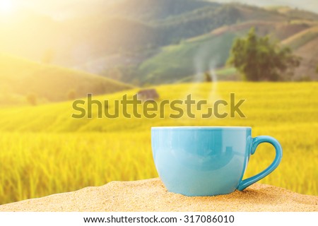 blue cup of coffee on white sand over village is surrounded by rice terraces on day noon light background.