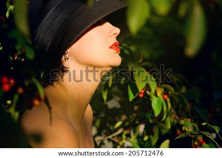 Head shod portrait of a beautiful woman in her forties sitting profile in black hat with her bare shoulders and neck in sun beat, green foliage, summer, autumn. Selective focus