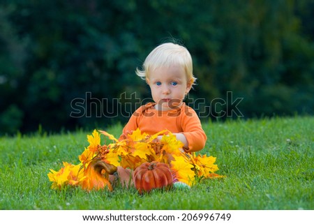 Portrait of a cute funny adorable blond Caucasian baby toddler with blue eyes in orange shirt sitting in the grass on the field meadow with yellow autumn fall leaves with pumpkins. Halloween, holidays