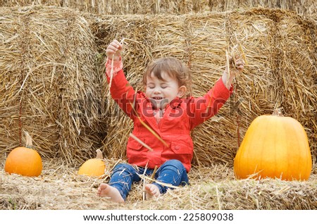 Portrait of a cute funny adorable smiling Caucasian baby toddler in red jacket and blue jeans sitting on the haystack on farm with pumpkins and playing with hay. Halloween Thanksgiving card