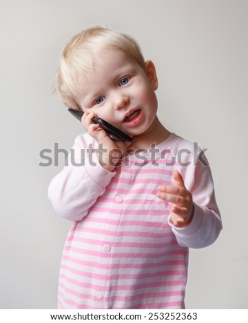Cute adorable white  blond baby with blue eyes talking expressively emotionally over mobile cell phone with funny expression on her face, new technology future generation concept, on light background