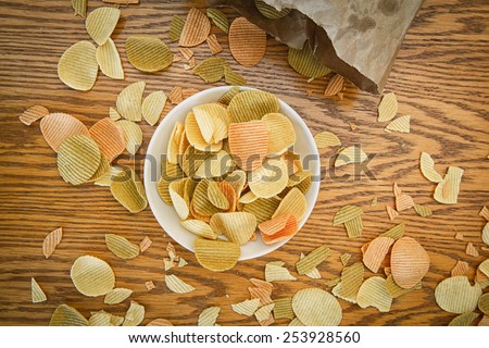 Closeup view from above of colorful veggie potato, tomato, spinach chips on a white plate and brown paper bag with greasy spots stains, on a wooden table