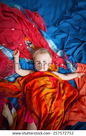 Magical cute blond Caucasian baby girl in skirt laying on a blue bed sheet covered with beautiful red, yellow, orange shawl cover. Concept childhood, happiness, fairy tale. View from above. Studio.