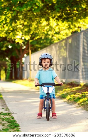 Portrait of a little boy toddler riding a balance bike in helmet on the road outside outdoors on a bright sunny spring summer day, seasonal child activity concept, healthy lifestyle