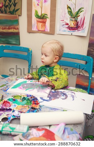 Cute adorable little baby boy girl toddler sitting in art studio indoors and drawing painting with brushes, pencils, pastel. Early child development concept
