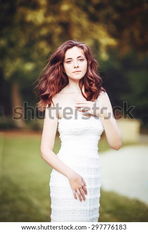 Portrait of beautiful  innocent Caucasian adult girl woman with long dark red brown hair, hazel eyes in white summer open dress, her hands arms on her chest standing in park outside looking in camera