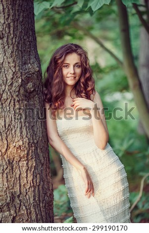 Portrait of beautiful innocent Caucasian adult girl woman with long dark red brown hair, hazel eyes in white summer open dress, her hands arms on her chest standing in park outside looking in camera