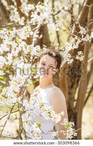 Closeup portrait of beautiful sexy Caucasian adult girl woman with dark brown hair and hazel eyes in white dress looking in camera among blooming trees sakura flowers in spring summer