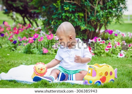 Portrait of cute adorable little indian South Asian infant boy in white shirt sitting on ground with toys in park green grass blanket outside on bright summer day smiling and playing