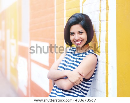 Portrait of beautiful smiling young hipster latin hispanic girl woman with short hair bob, in blue white striped tshirt, leaning on brick wall in city looking in camera, lifestyle fashion concept