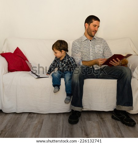 Baby playing with tablet and father reading a book on a white sofa both without care of each other with red pillow and little bear