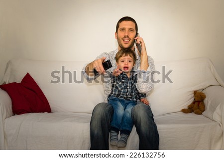 Baby and father watching tv on a white sofa while baby is crying and father is making a phone call, with red pillow and little bear
