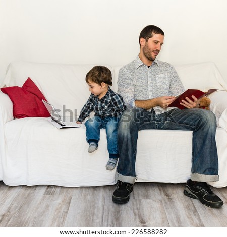 Baby playing with tablet and father reading a book on a white sofa