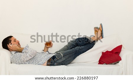Concentrated young man lying on the couch drinking and making a phone call on a white sofa with a red pillow