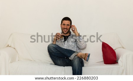 Young man drinking and making a phone call smiling on a white sofa with a red pillow