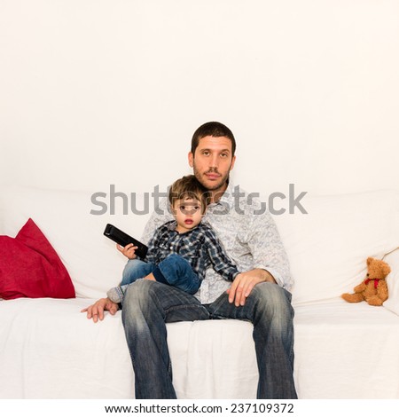 Bored father watching tv with his son sitting on his knees