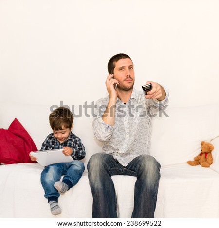 Bored father making a phone call and watching tv while baby is playing with tablet on a white sofa with a red pillow and a bear toy