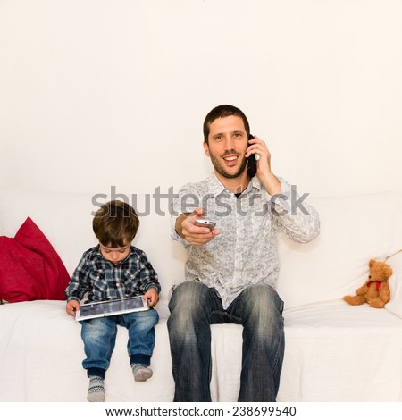 Smiling father making a joke with phone and tv remote control while son is playing with tablet on a white sofa with a red pillow and a bear toy