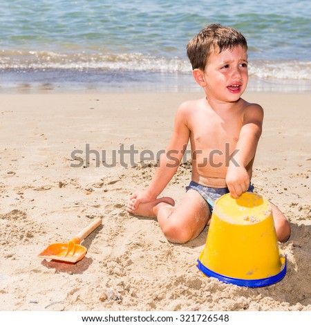 cute caucasian child crying while playing on the beach with pail and shovel in a sunny day with sea background