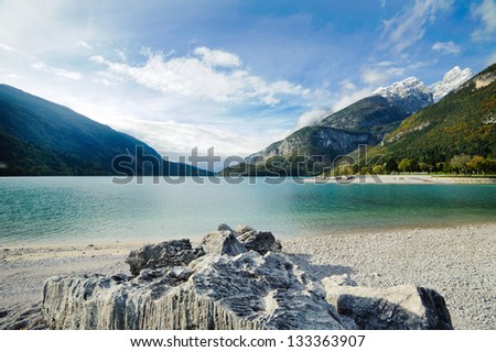 Lake with clear clean water on a background of mountains.  Blue sky with light clouds.  The lake water is turquoise and clear. There are big grey stones in the foreground.