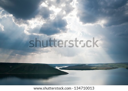 Rays of sun shining through the clouds. The river runs into the distance. The river is calm. The sky is cloudy.