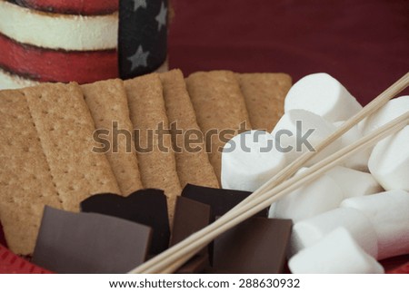 S\'more ingredients, on a red tablecloth, with an american flag, painted, candle in the imediate background.