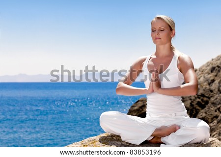 Beautiful positiveblond girl clothing in white sit at the seaside on the rock and meditating in yoga pose - stock photo