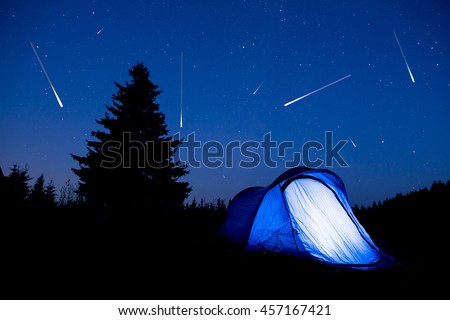 Blue illuminated tent with travelers in the mountain. Background of a pine tree silhouette and the starry summer night sky. Meteor shower. Falling stars.