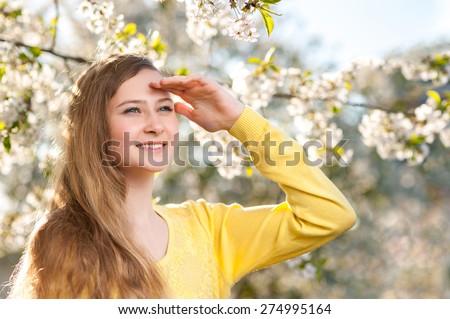 A young smiling girl looks ahead far and holds a hand above eyes. Spring cherry trees with white flowers on a background.