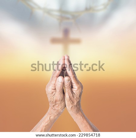 Hands of old human praying over blurred crown of thorns and the cross on a sunset.