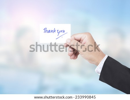 Asian business man holding a handwritten THANK YOU over blurred working people in office background.