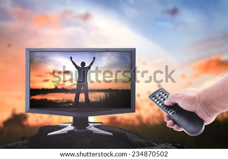Human hands holding remote and monitor display silhouette of man with hands raised to beautiful sunset over the actual location.
