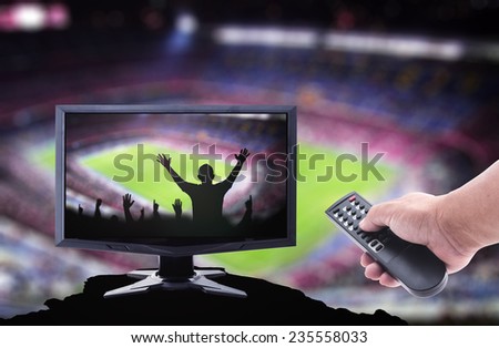 Human hands holding remote and monitor display Silhouettes people raising hands over stadium on the actual location.