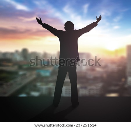 Silhouette businessman raising hands over blurred city on sunset background.