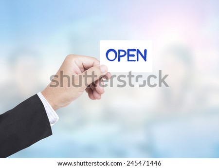 Asian business man holding a handwritten OPEN over blurred working people in office background.