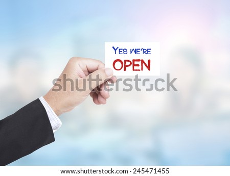 Asian business man holding a handwritten YES WE\'RE OPEN over blurred working people in office background.
