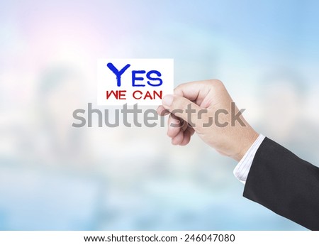 Asian business man holding a handwritten YES WE CAN over blurred working people in office background.