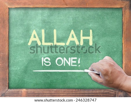 Human hand holding chalk and writing text for ALLAH IS ONE! on green board.
