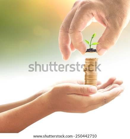 Human hand adding a golden coin with young plant into golden coins in another hands on blurred sunset background. Money coin concept.
