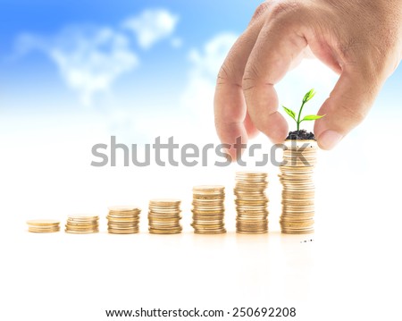 Human hand adding a golden coin with young plant in the final row over blurred world map of clouds background. Money coin concept.