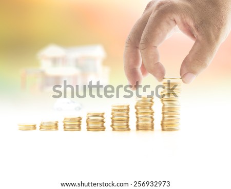 Human hand adding a golden coin in the final row of golden coins over blurred house and car on sunset background. Concept for money coin, insurance, buying, renting, service.
