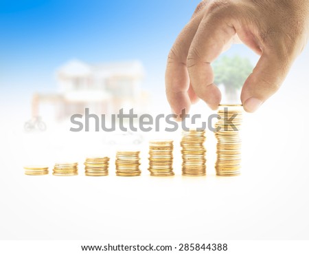 Human hand adding a golden coin in the final row of golden coins over blurred house and car on blue sky background. Concept for money coin, insurance, buying, renting, service.