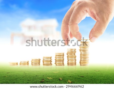 Human hand adding a golden coin in the final row of golden coins on meadow over blurred house on blue sky background. Concept for money coin, insurance, buying, renting, service.